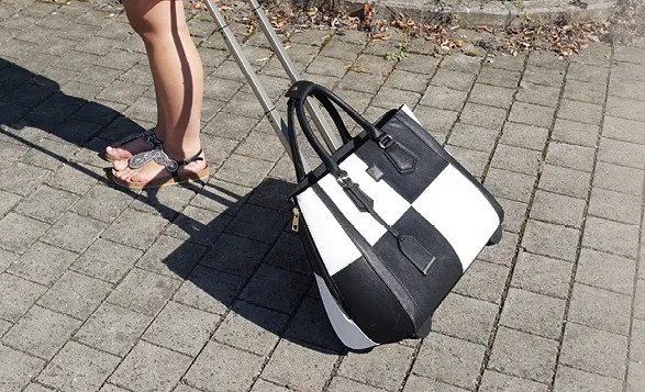 Frau zieht einen schwarz-weiß karierten Trolley über Pflastersteinweg. 