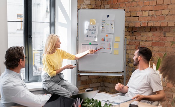 Frau präsentiert vor Whiteboard mit Diagrammen, zwei Kollegen hören zu.